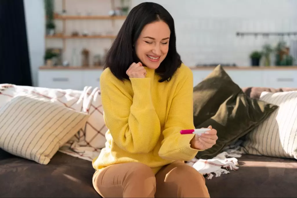 жена с черна коса и жълт пуловердържи тест за бременност