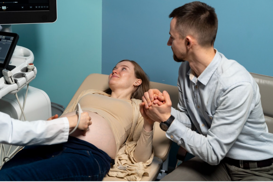 Бременна жена, придружена от партньора си е на преглед с ехограф в лекарски кабинет.