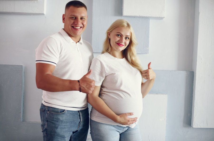 Усмихната млада двойка - мъж и бременна жена с вдигнати нагоре палци на ръцете.