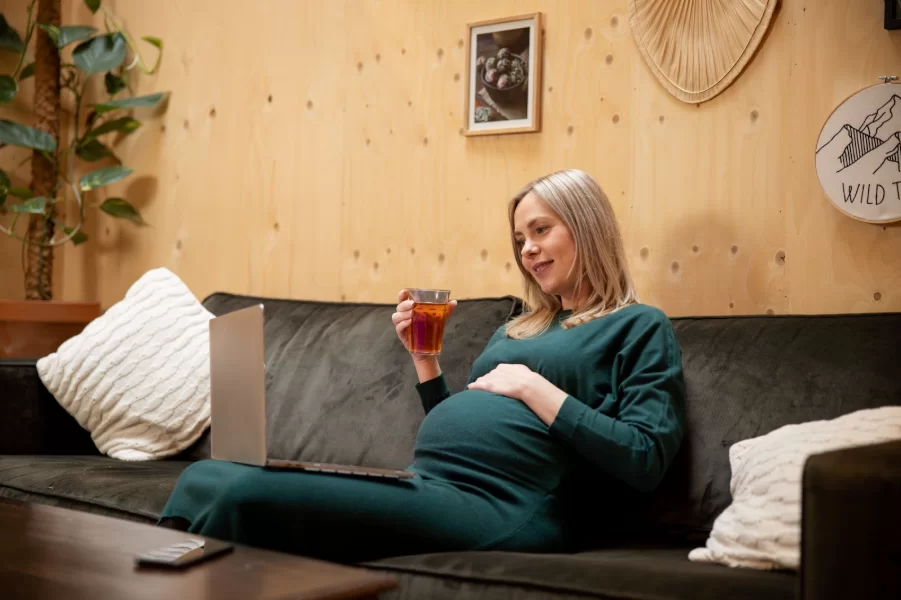 Бременна жена в зелена рокля е седнала на диван, гледа в екрана на лаптоп и държи напитка в ръка.